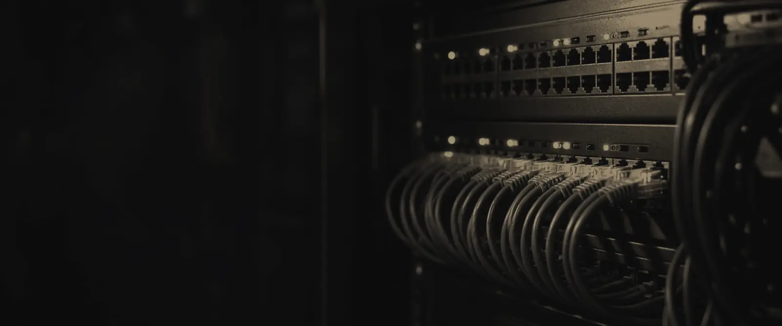 A network patch panel in a dim equipment closet, rows of cables neatly run between ports, with small status lights on.